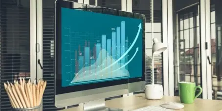 a desk with a cup full of pencils, a green mug and a computer screen that is displaying a blue bar graph with numbers and upward arrows