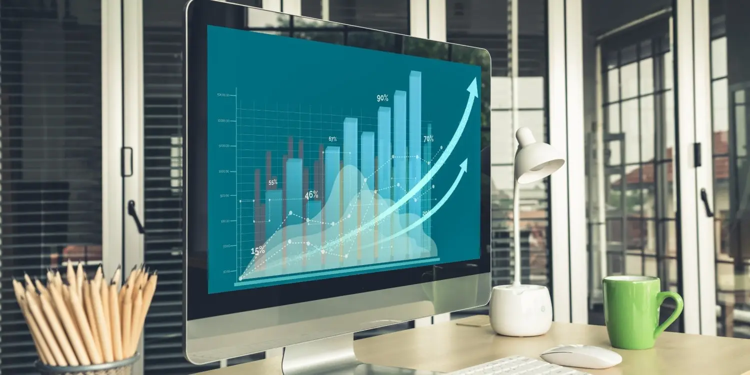 a desk with a cup full of pencils, a green mug and a computer screen that is displaying a blue bar graph with numbers and upward arrows
