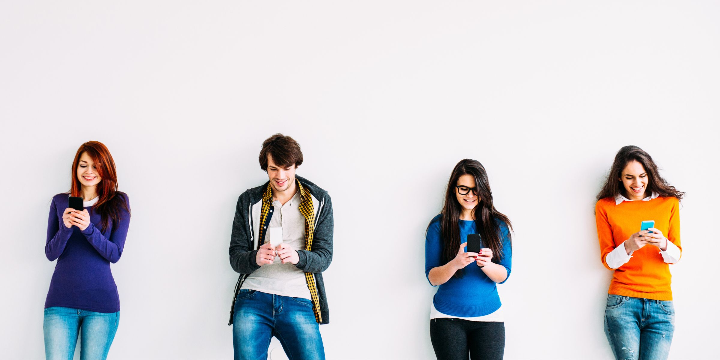 four students standing in front of a white background and smiling at their cell phones 