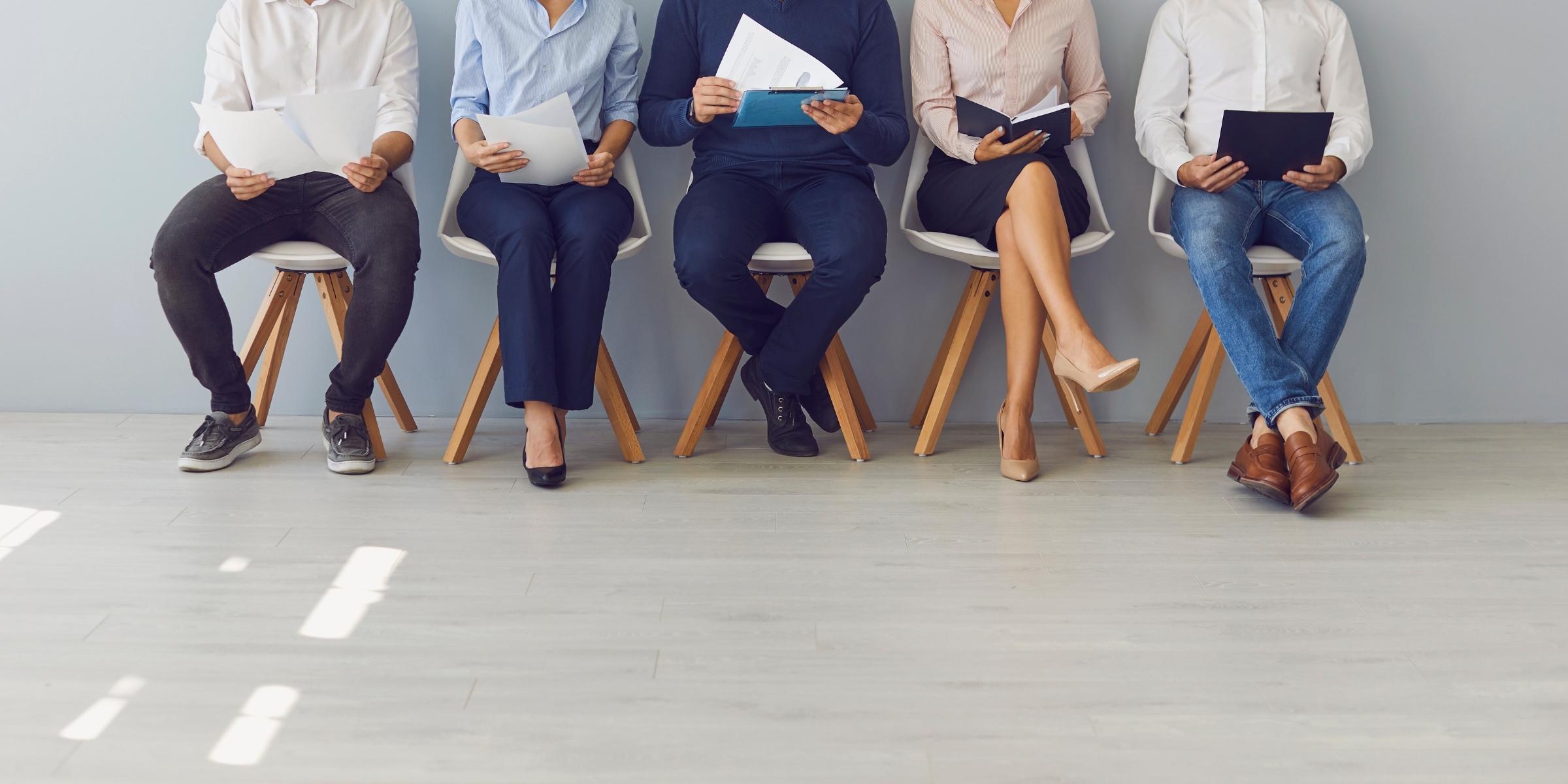 closeup of five people in business casual clothing sitting close to each other on chairs and holding slips of paper, likely their resumes