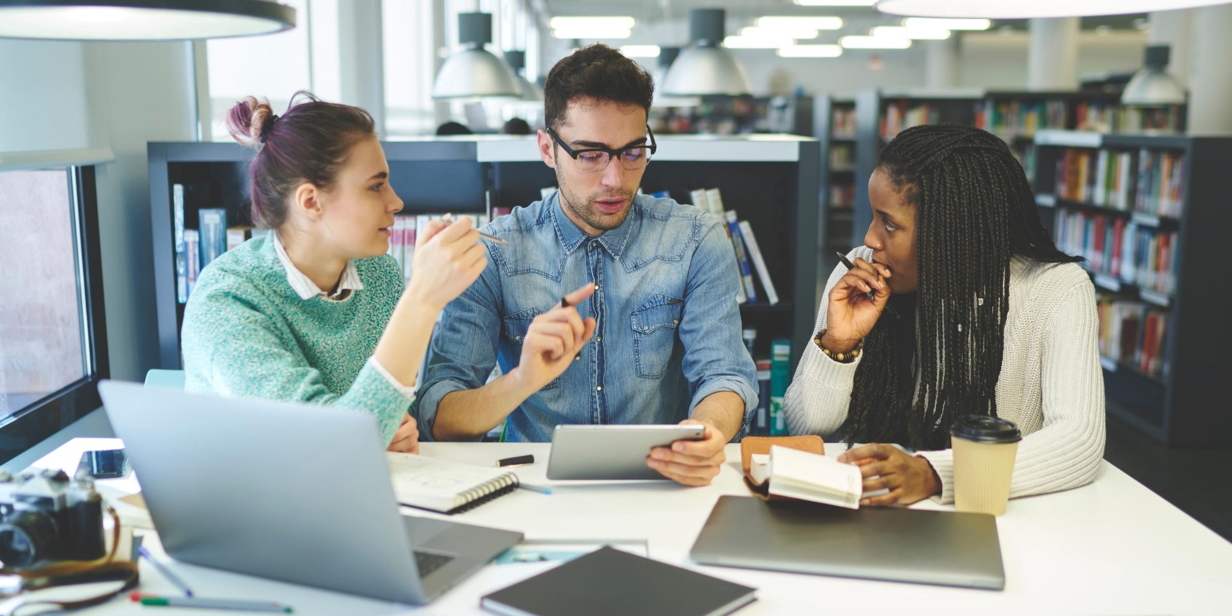three students chatting around a table with laptops and tablets