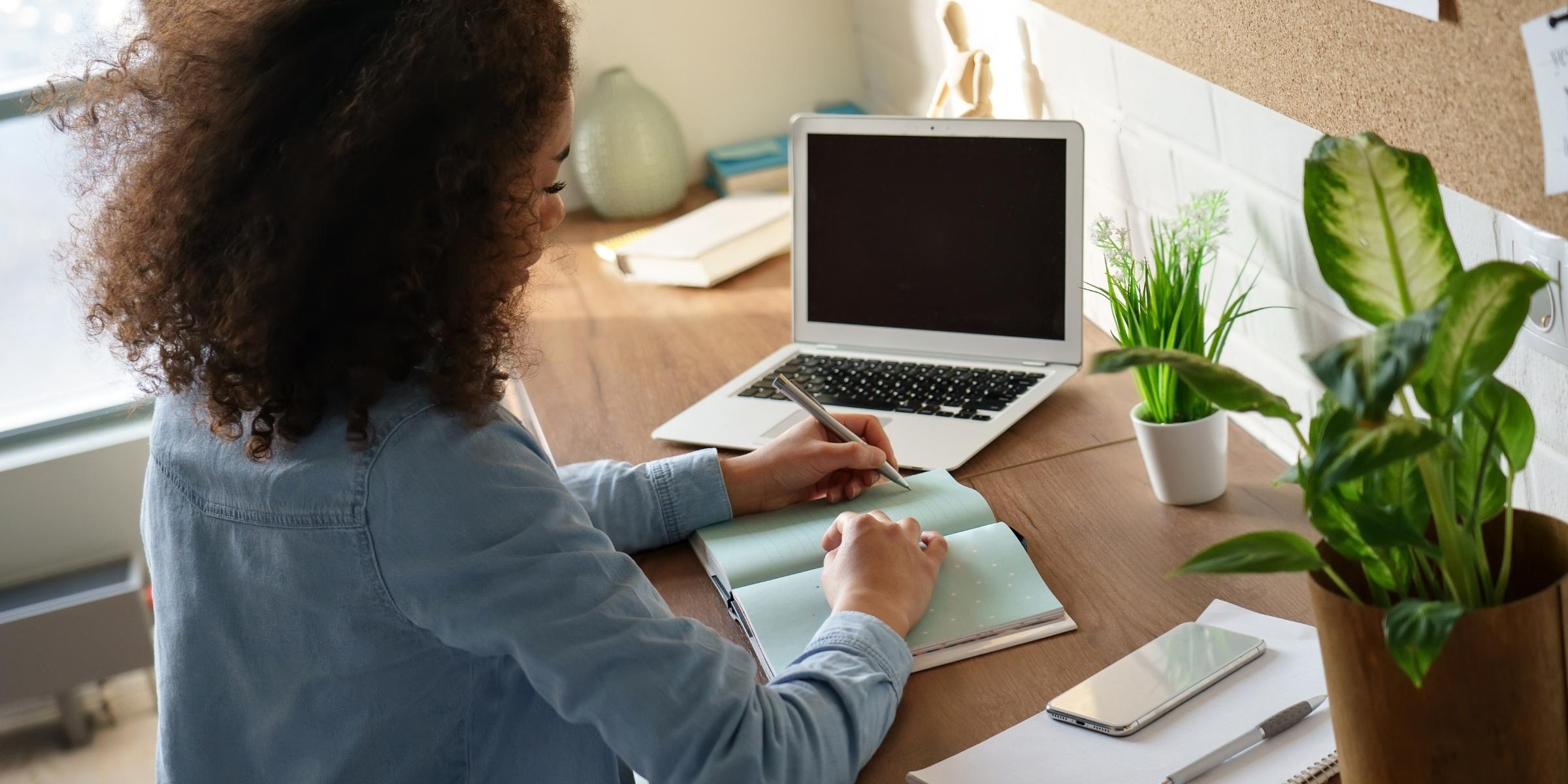 a person with tall, curly hair writing in a notepad next to a laptop